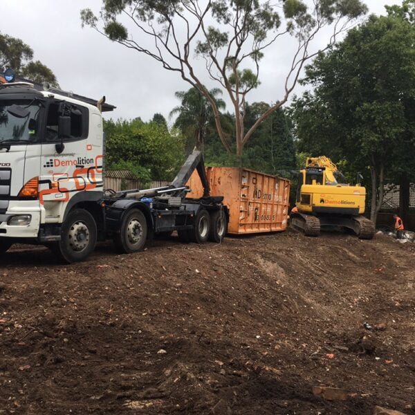 A truck and an excavator from a home demolitions company are parked at a construction site with a large dumpster in the background. Trees and debris are visible around the site.