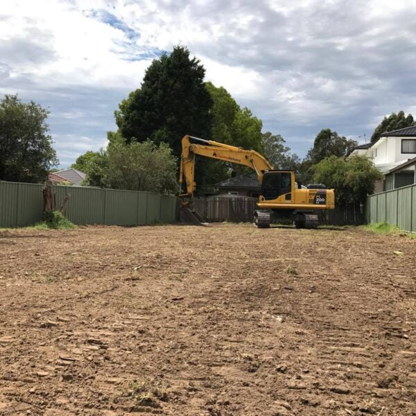 A yellow excavator, ready for home demolitions, sits idle in the middle of a large, cleared dirt lot surrounded by green metal fencing, with trees and residential buildings in the background under a cloudy sky.