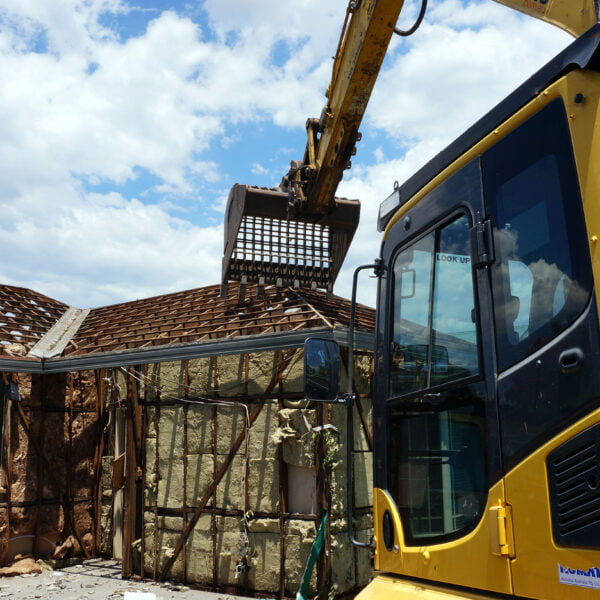 A large yellow excavator is taking center stage in home demolitions, tearing through a partially deconstructed house under a cloudy sky. The house's wooden frame and insulation are exposed to the elements.