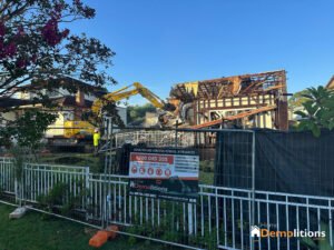 A demolition site with an excavator tearing down a house surrounded by a fence. A sign by the entrance reads “Home Demolitions - Demolition and asbestos removal in progress.” The site, once an art gallery, now witnesses its walls crumbling to dust.