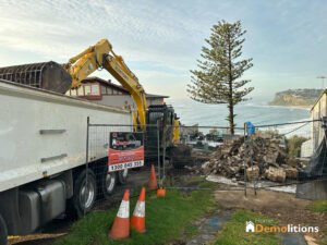 An excavator loads debris into a truck at a coastal demolition site enclosed by a fence labeled "Home Demolitions." Bricks and rubble are piled on the ground, much like an open-air gallery of destruction; safety cones are placed nearby.