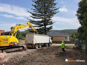 A yellow excavator loads debris into a white truck at a construction site. Nearby, a worker in a green vest and white helmet stands attentively. The scene, framed by trees and a house in the background, resembles an industrial exhibition of precision and teamwork.