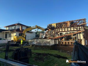 An excavator demolishes a house while a person in a high-visibility vest supervises. The yard is fenced, showcasing a sign for Home Demolitions in the foreground, resembling an industrial gallery of destruction.