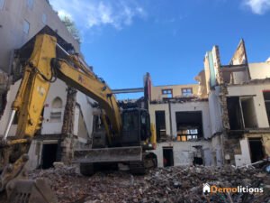 A large yellow excavator is demolishing a multi-story building, creating a scene that could be mistaken for an art exhibition. Debris and rubble are scattered around the site, resembling an unconventional gallery against the backdrop of a clear blue sky contrasting with the destruction.
