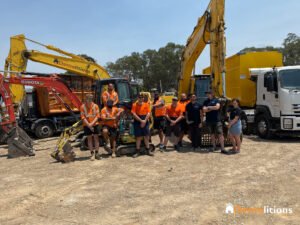A group of construction workers stands in front of yellow excavators and heavy machinery in a dirt lot, reminiscent of an industrial gallery. They are wearing orange safety vests, with trees visible in the background.