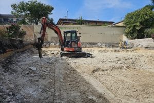 An excavator digs at a construction site with surrounding walls and trees under a clear sky.