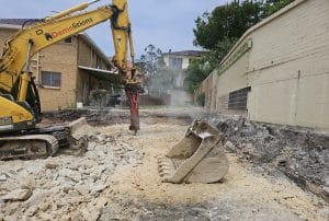 Excavator breaking ground with a hydraulic hammer in an urban area, surrounded by walls and buildings. Dust is visible in the air.