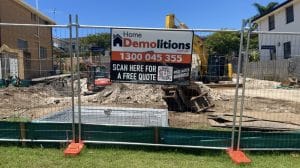 A demolition site featuring a fence with a "Home Demolitions" sign stands ready, while construction machinery looms in the background. Measures for sediment control ensure the area remains secure and environmentally responsible amidst the ongoing work.