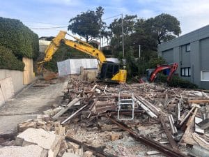 The demolition site, with debris and two excavators—one yellow and one red—amidst fallen wooden and concrete materials, features effective sediment control measures. Trees and a modern building provide a contrasting backdrop to the construction chaos.