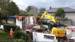 A yellow excavator efficiently demolishes older homes while a worker in an orange outfit sprays water nearby. In the background, a gray-roofed building, surrounded by trees, awaits its transformation into an energy-efficient space.