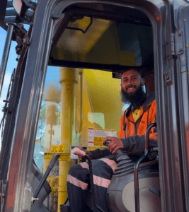 A person in a high-visibility jacket sits inside a construction vehicle, smiling while holding a lever, embodying the next chapter of Nick's Journey as a skilled demolition operator.