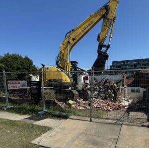 An excavator from a renowned demolition service is safely demolishing a brick building behind a fenced area. Debris scatters the scene, and a diligent worker in a helmet stands nearby, ensuring all safety measures are followed for efficient home demolitions.