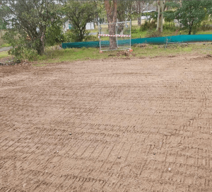 A leveled dirt area with visible tracks indicates the initial construction steps, surrounded by trees and a construction barrier in the background.