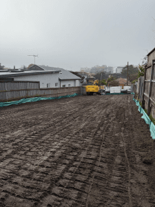 A cleared and leveled construction site with a yellow excavator and green tarps along the fence on both sides. Houses and buildings are visible in the background.