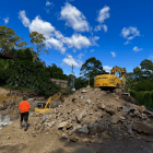 A construction worker walks toward two excavators on a dirt site, with a pile of rocks in the foreground and trees under a blue sky with clouds.
