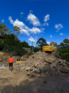 A construction worker walks toward two excavators on a dirt site, with a pile of rocks in the foreground and trees under a blue sky with clouds.