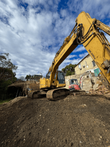 A yellow excavator sits on a mound of dirt at a construction site, with a smaller red excavator and residential buildings in the background under a partly cloudy sky.