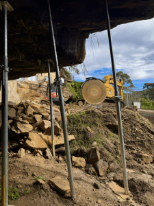 View from beneath an overhanging rock supported by scaffolding poles; a yellow excavator with a large circular saw is positioned on rocky ground outside.