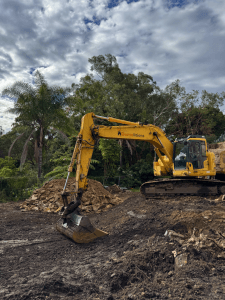 A yellow excavator is parked on a dirt construction site near piles of rocks and soil, surrounded by trees under a partly cloudy sky.