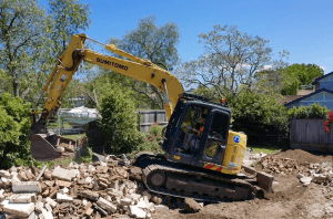 A yellow excavator moves debris and rubble at a demolition site, surrounded by trees and houses under a clear blue sky.
