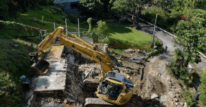 A yellow excavator demolishes a house surrounded by greenery, with debris scattered around and a dirt path nearby.