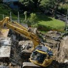 A yellow excavator demolishes a house surrounded by greenery, with debris scattered around and a dirt path nearby.