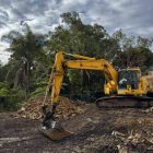 A yellow excavator is parked on a dirt construction site near piles of rocks and soil, surrounded by trees under a partly cloudy sky.