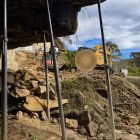 View from beneath an overhanging rock supported by scaffolding poles; a yellow excavator with a large circular saw is positioned on rocky ground outside.