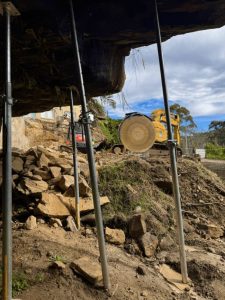 View from beneath an overhanging rock supported by scaffolding poles; a yellow excavator with a large circular saw is positioned on rocky ground outside.