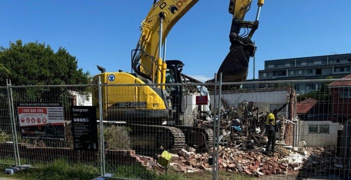 An excavator from a renowned demolition service is safely demolishing a brick building behind a fenced area. Debris scatters the scene, and a diligent worker in a helmet stands nearby, ensuring all safety measures are followed for efficient home demolitions.