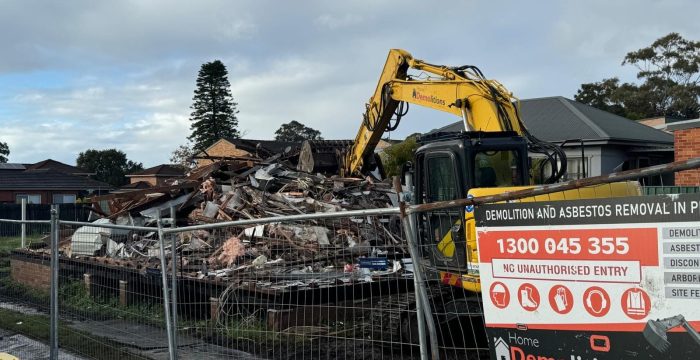 An excavator skillfully dismantles a house, surrounded by rubble and vital safety warnings on a metal fence. Contact information for the experienced demolition company is prominently displayed, ensuring the process is both safe and professional.