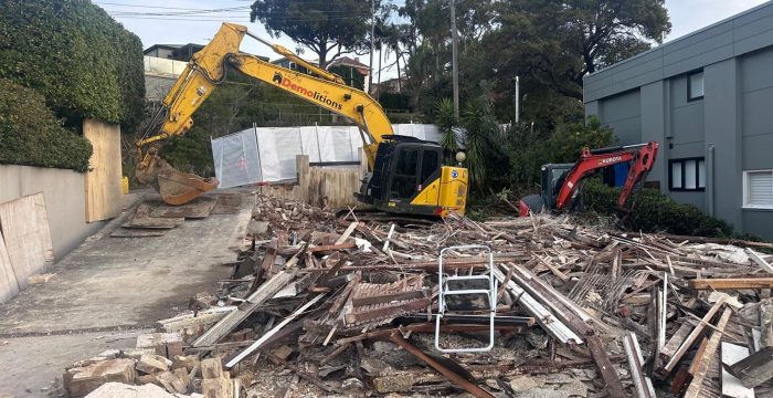The demolition site, with debris and two excavators—one yellow and one red—amidst fallen wooden and concrete materials, features effective sediment control measures. Trees and a modern building provide a contrasting backdrop to the construction chaos.