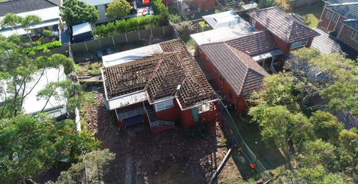 Aerial view of a partially demolished red-brick house with exposed roof beams and debris surrounding the property, situated between two intact houses—a stark reminder of ongoing home demolitions in the area.