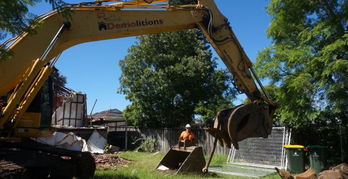 A large yellow excavator with the branding "Home Demolitions" is seen in a grassy yard, operated by a person in an orange vest and helmet. Debris and metal fencing are visible in the background.