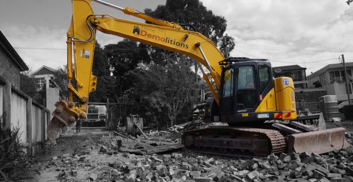 A yellow excavator with "Home Demolitions" on the arm is parked on a pile of rubble at a construction site surrounded by trees and buildings.