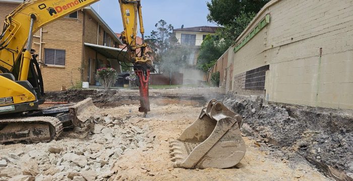 Excavator breaking ground with a hydraulic hammer in an urban area, surrounded by walls and buildings. Dust is visible in the air.