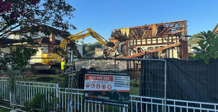 A demolition site with an excavator tearing down a house surrounded by a fence. A sign by the entrance reads “Home Demolitions - Demolition and asbestos removal in progress.” The site, once an art gallery, now witnesses its walls crumbling to dust.
