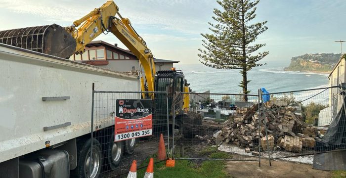An excavator loads debris into a truck at a coastal demolition site enclosed by a fence labeled "Home Demolitions." Bricks and rubble are piled on the ground, much like an open-air gallery of destruction; safety cones are placed nearby.