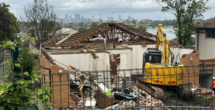 A partially demolished house with a visible excavator, set against a backdrop of a city skyline. Trees and bushes surround the site, and a fence is in the foreground, resembling an open-air gallery of urban transformation.