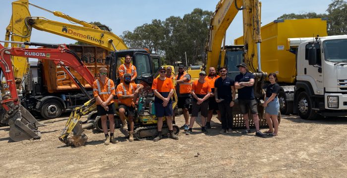 A group of construction workers stands in front of yellow excavators and heavy machinery in a dirt lot, reminiscent of an industrial gallery. They are wearing orange safety vests, with trees visible in the background.