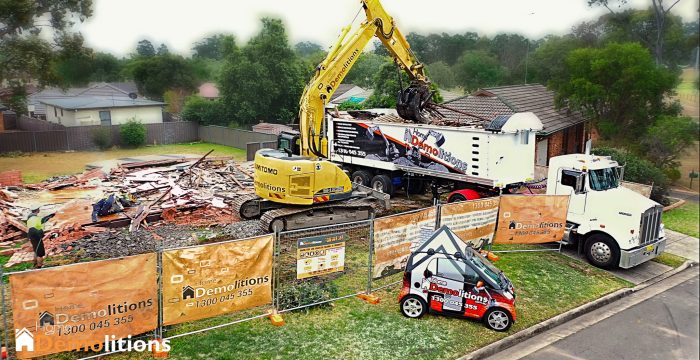 Heavy machinery is demolishing a house, with construction barriers and company signs visible. A truck and a small promotional vehicle for a nearby art gallery are parked nearby, hinting at an upcoming fine art exhibition.