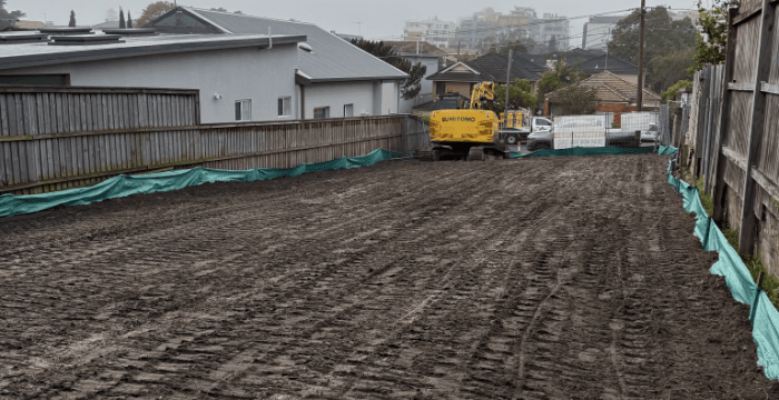 A cleared and leveled construction site with a yellow excavator and green tarps along the fence on both sides. Houses and buildings are visible in the background.