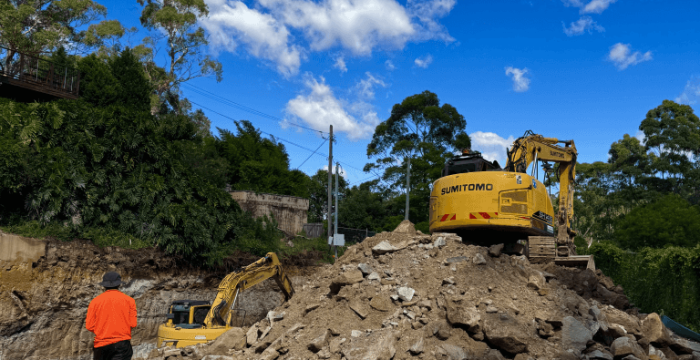 A construction worker walks toward two excavators on a dirt site, with a pile of rocks in the foreground and trees under a blue sky with clouds.