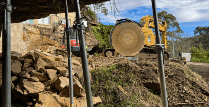 View from beneath an overhanging rock supported by scaffolding poles; a yellow excavator with a large circular saw is positioned on rocky ground outside.