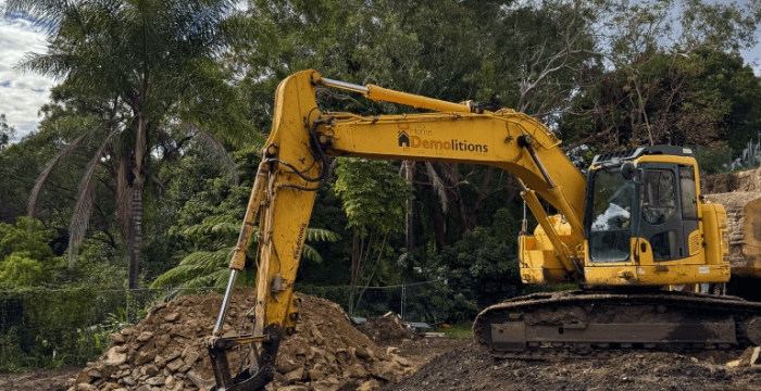 A yellow excavator is parked on a dirt construction site near piles of rocks and soil, surrounded by trees under a partly cloudy sky.