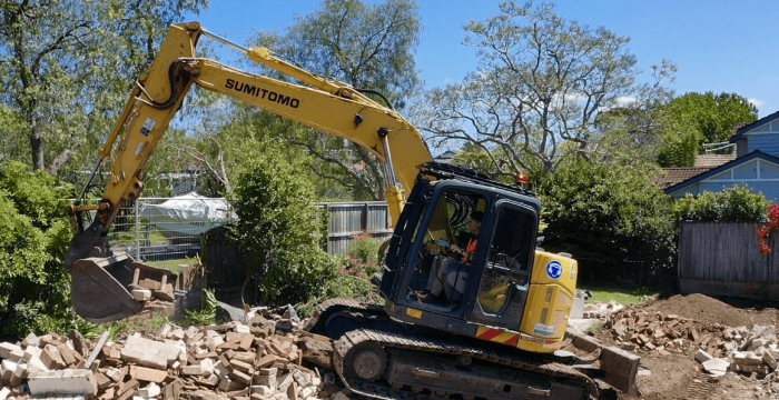 A yellow excavator moves debris and rubble at a demolition site, surrounded by trees and houses under a clear blue sky.