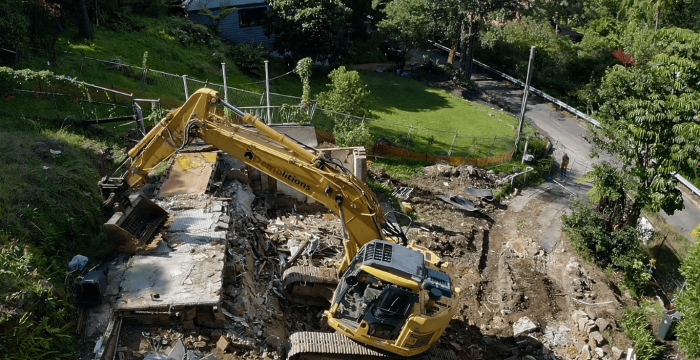 A yellow excavator demolishes a house surrounded by greenery, with debris scattered around and a dirt path nearby.