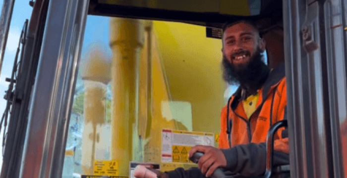A person in a high-visibility jacket sits inside a construction vehicle, smiling while holding a lever, embodying the next chapter of Nick's Journey as a skilled demolition operator.