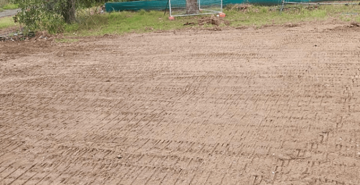 A leveled dirt area with visible tracks indicates the initial construction steps, surrounded by trees and a construction barrier in the background.