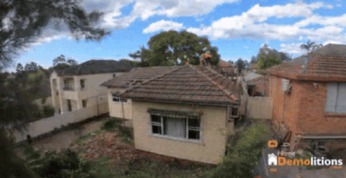 A house is being demolished, with one worker on the roof and debris falling. The Home Demolitions logo is visible in the bottom right corner, reminding onlookers of their collaboration with New Homebuilders Grant Australia.
